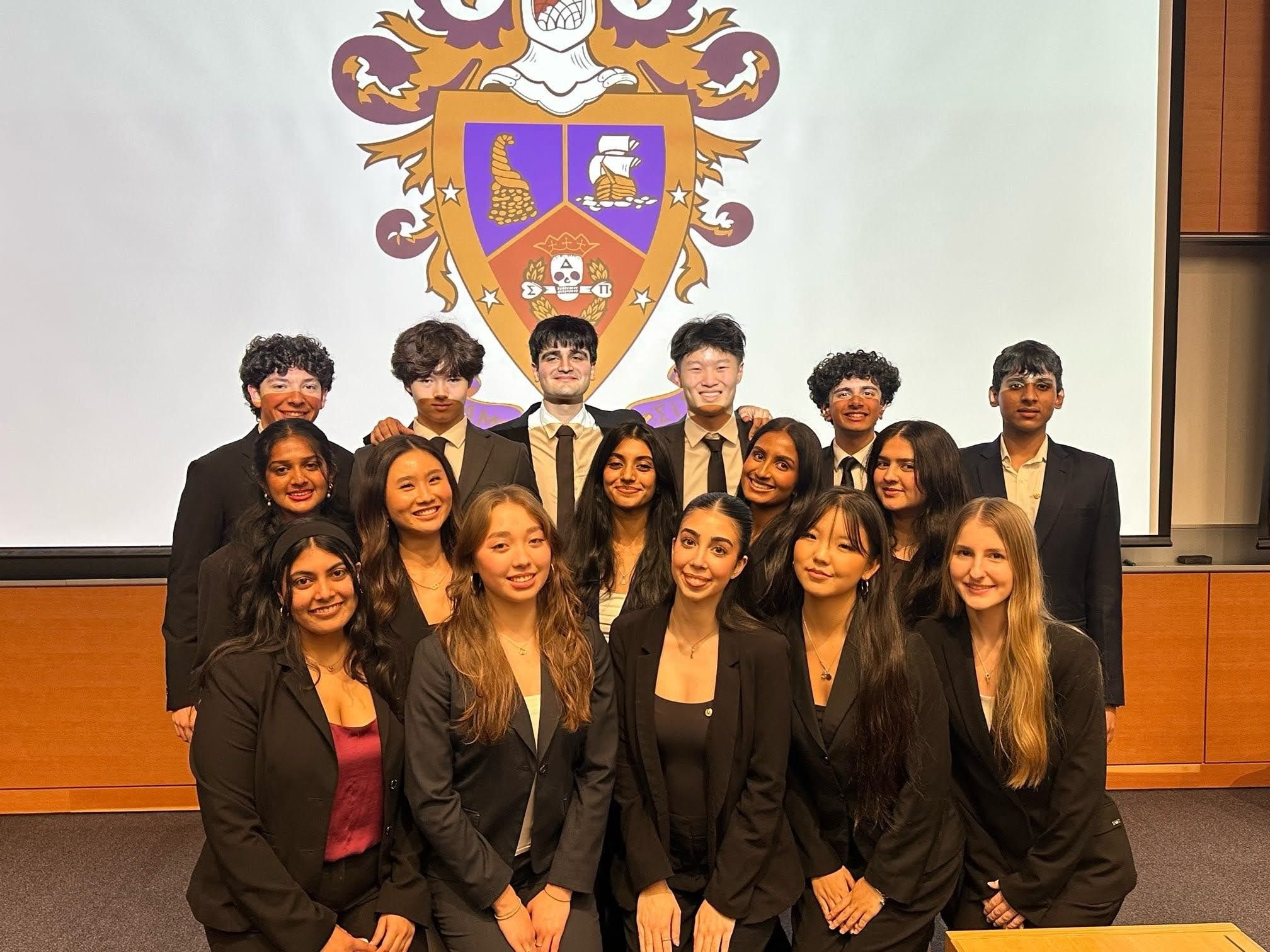 Brothers in business attire in front of the Delta Sigma Pi crest projected on a lecture hall screen