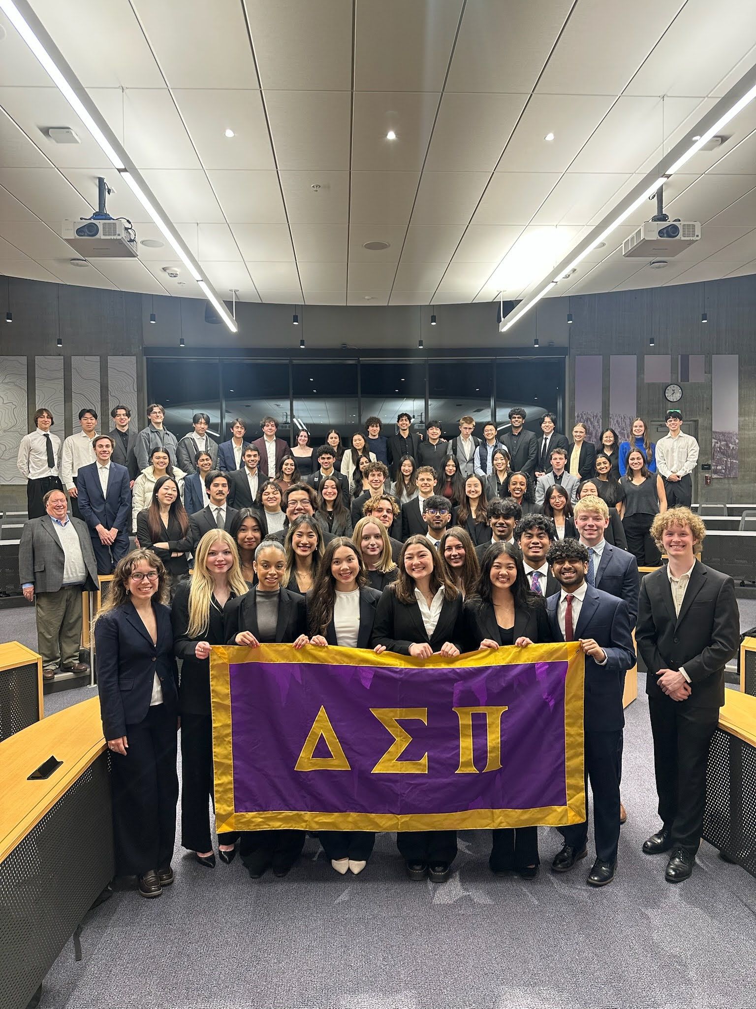Sigma Upsilon brothers in business attire filling a lecture hall, with the front row holding a purple and gold Delta Sigma Pi banner